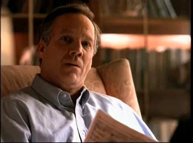 A middle-aged man in a light blue shirt sits in an armchair, holding a newspaper, looking thoughtful in a cozy interior.
