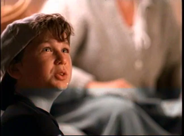 A young boy in a cap looks up and speaks, with an older person out of focus in the background, in an interior setting.