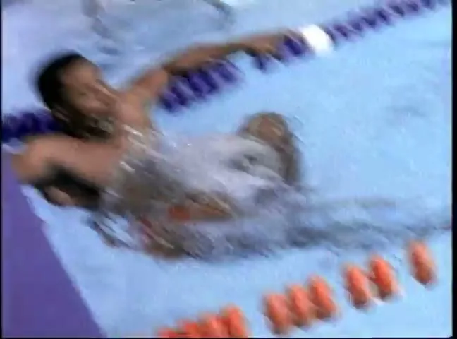 A male swimmer with dark hair swims freestyle in a pool, creating splashes, with lane lines visible in the foreground and background.
