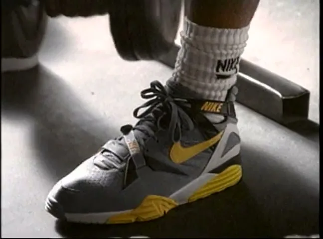 A person's foot wearing a grey and yellow Nike athletic shoe and white Nike socks is shown in a gym setting.