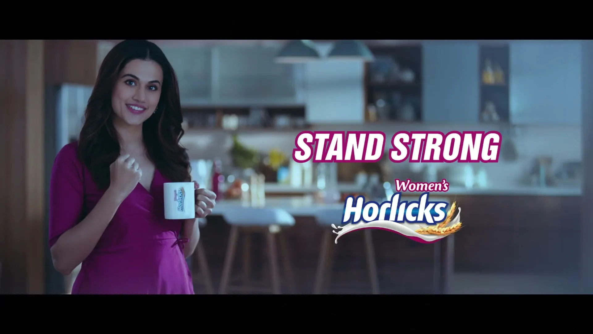 A young woman in a purple dress smiles at the camera while holding a mug in a modern kitchen, with product text on screen.