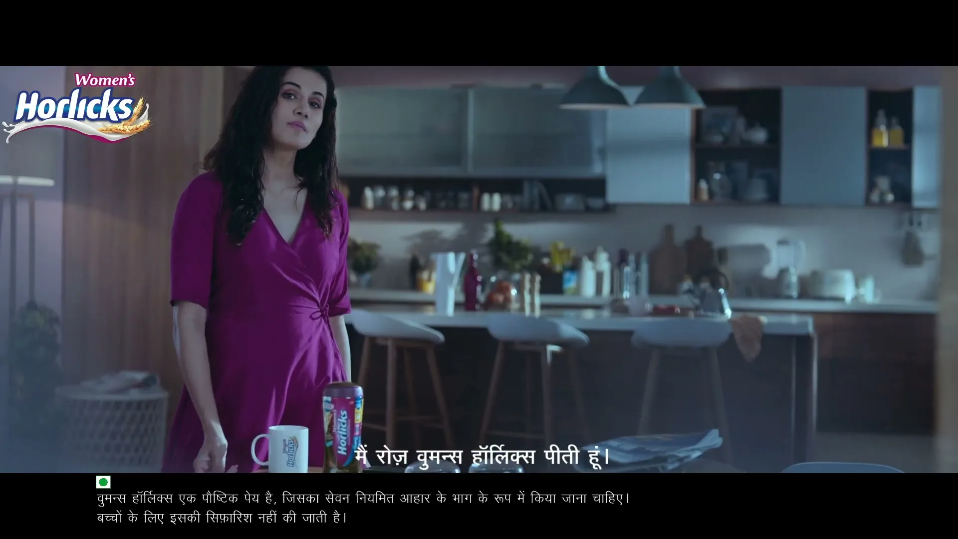 A woman in a purple dress stands in a modern kitchen, holding a spoon and looking at the camera.