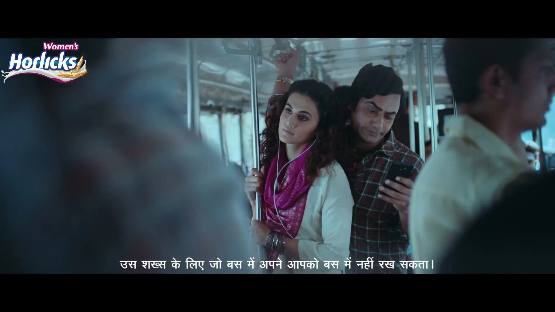 A young woman with curly hair and a pink scarf stands on a crowded bus, looking uncomfortable as a man behind her looks at his phone.