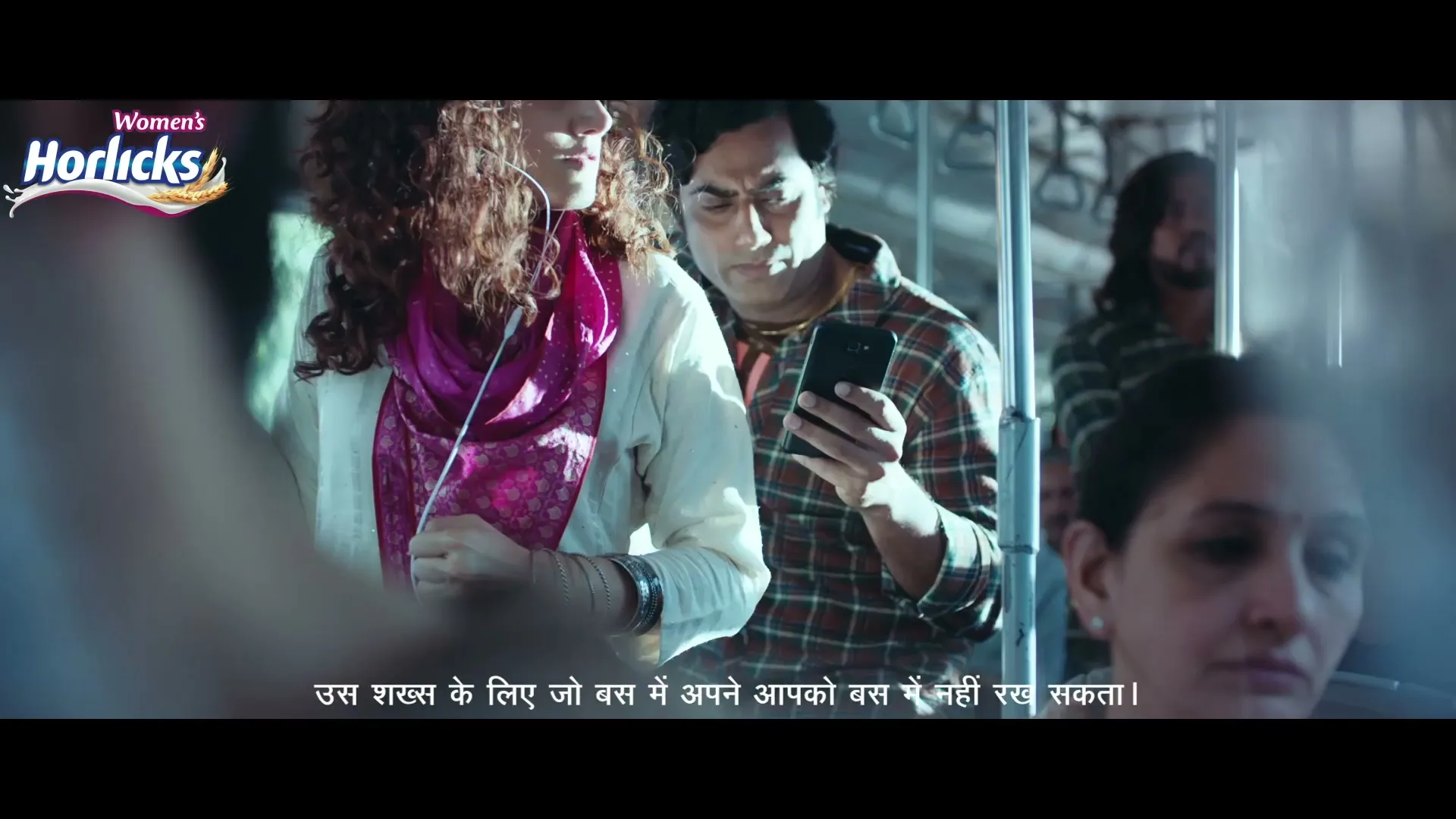 A young woman with curly hair and a pink scarf listens to music on a crowded bus, while a man next to her looks at his phone.