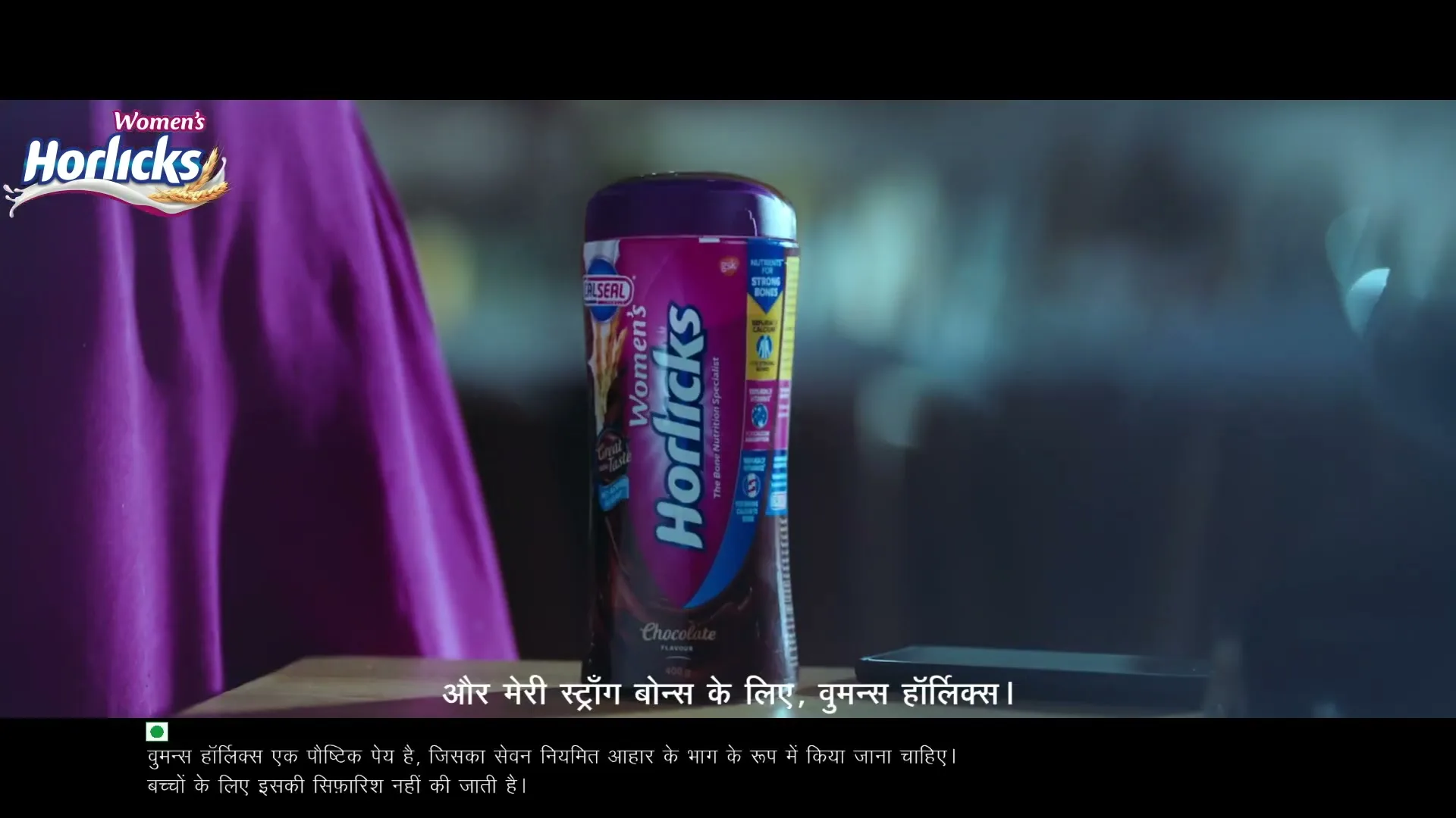 A purple Horlicks bottle stands on a wooden surface, with a woman in a purple sari partially visible in the background, in an interior setting.