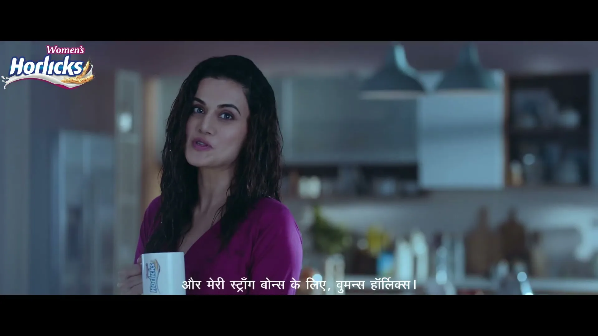 A woman in her 30s, with dark hair and a purple top, holds a white mug and smiles at the camera in a modern kitchen.