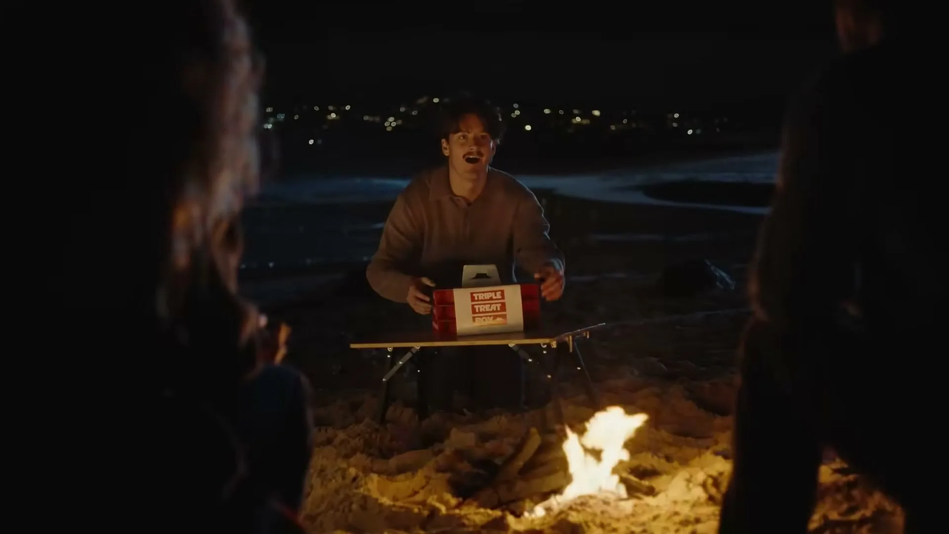 A young man sits by a campfire at night, holding a Pizza Hut Triple Treat Box, with two friends partially visible on either side.