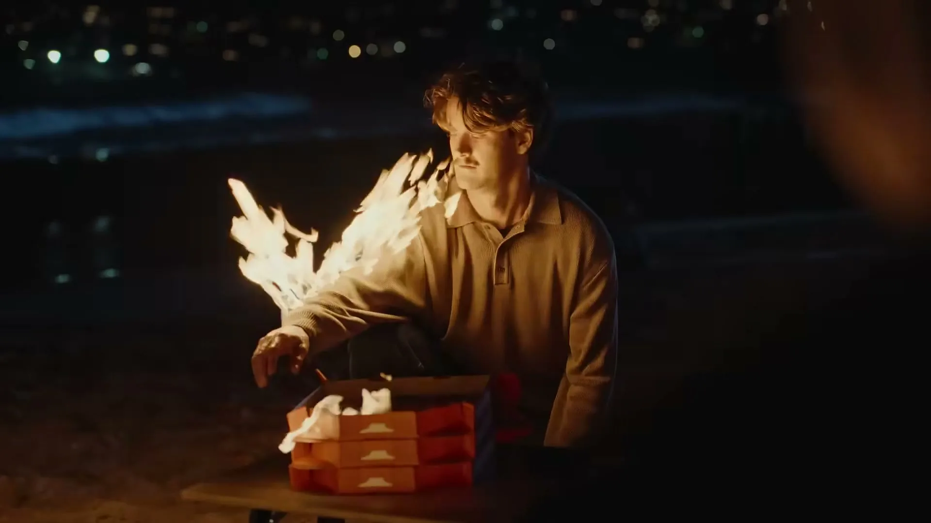 A young man with brown hair and a polo shirt sits on a beach at night, looking at a stack of pizza boxes that are on fire.