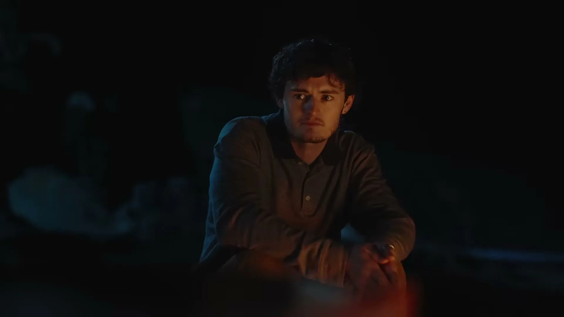 A young man with curly hair sits by a campfire at night, looking directly at the camera with a slight smile.