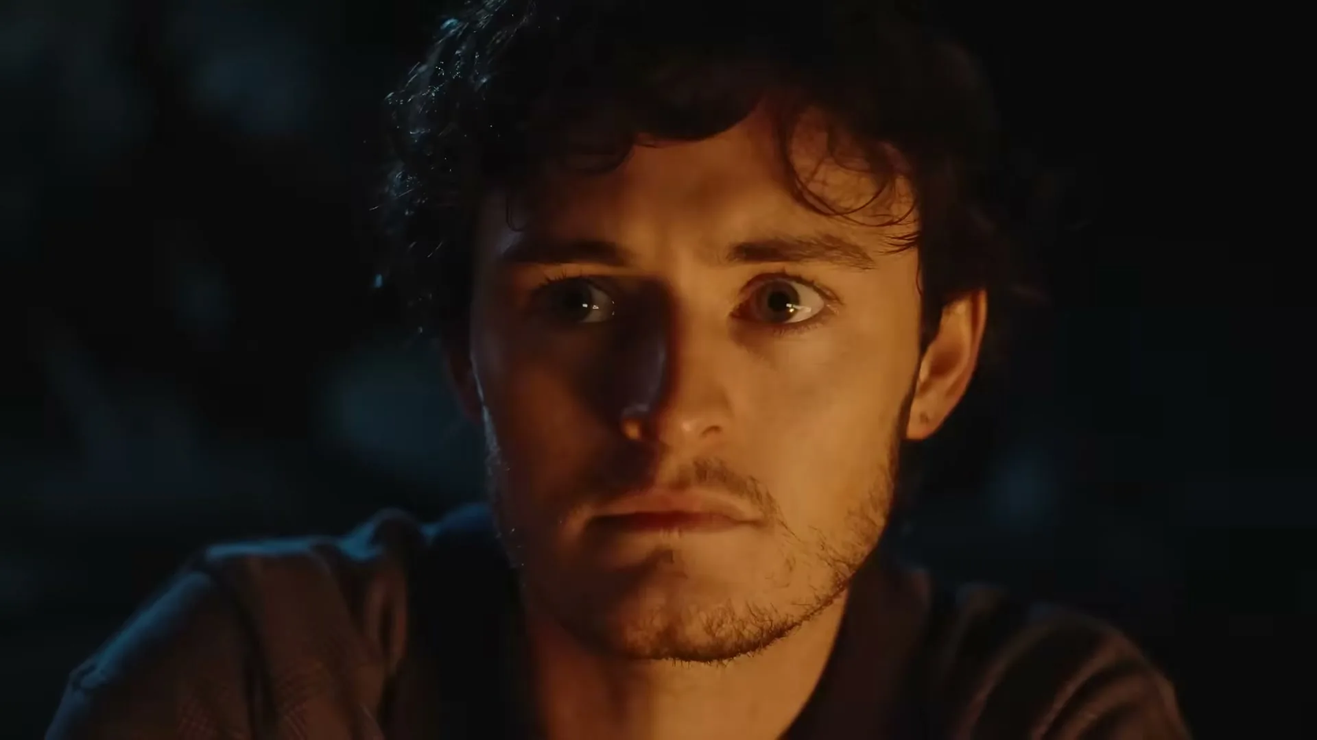 A young man with curly hair and a beard looks intently at something off-screen, illuminated by a warm glow in a dark setting.