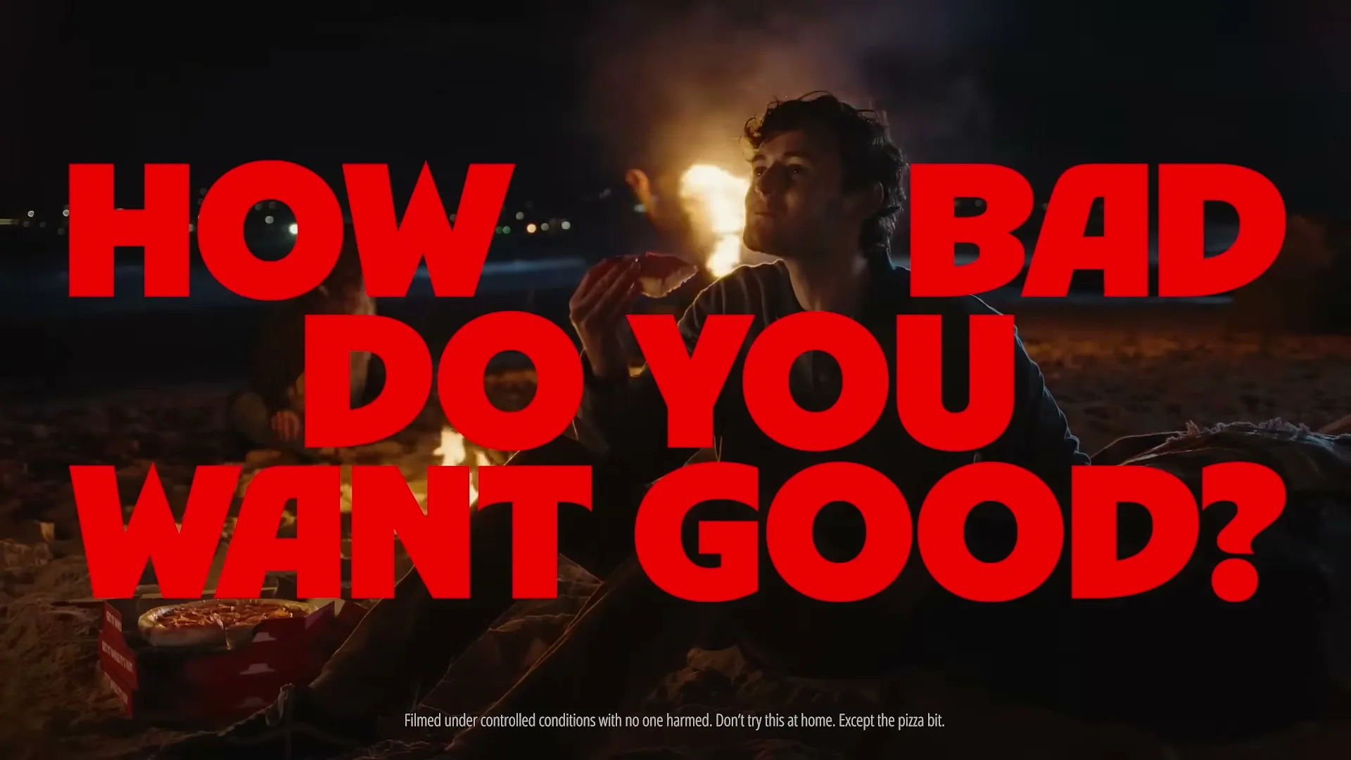 A young man sits on a beach at night, eating pizza by a campfire, with large red text overlayed on the screen.