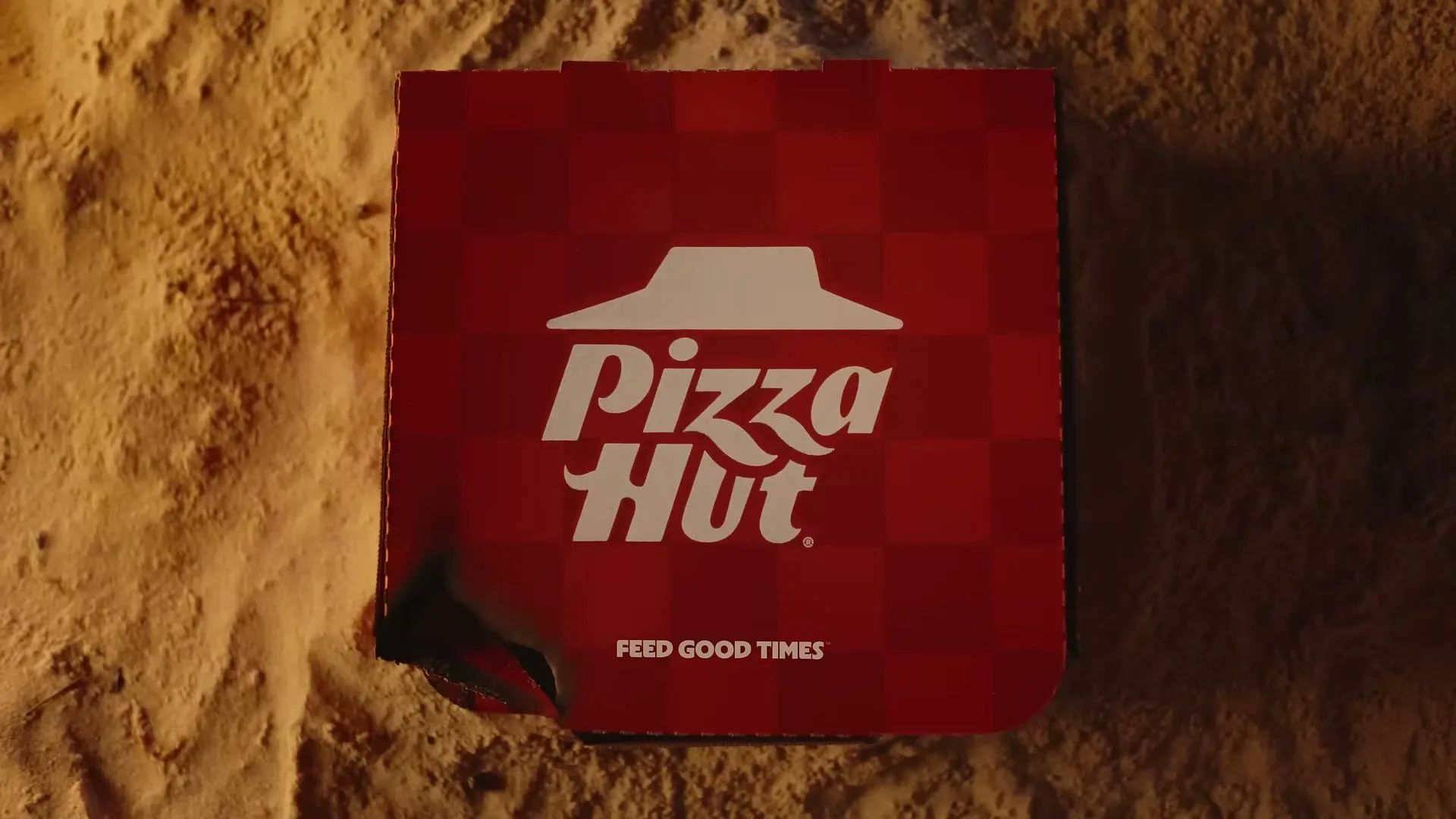 A red Pizza Hut pizza box with a white logo is seen from an overhead top-down angle, resting on a sandy surface.