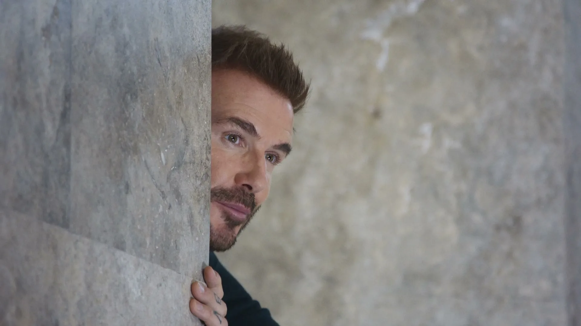 David Beckham peeks around a stone wall, looking to the right with a slightly mischievous expression in an interior setting.