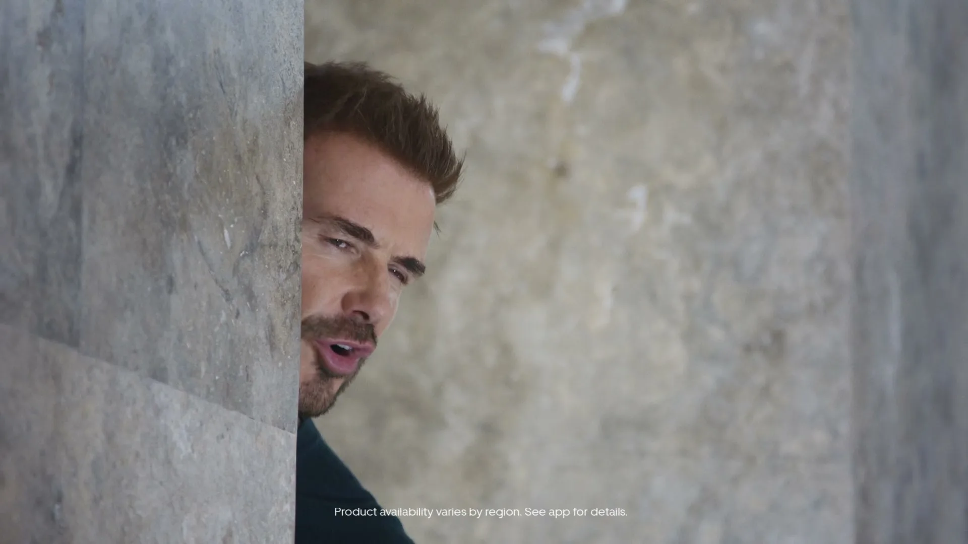A man with reddish-brown hair and a beard peeks from behind a stone wall, looking around with a curious expression.