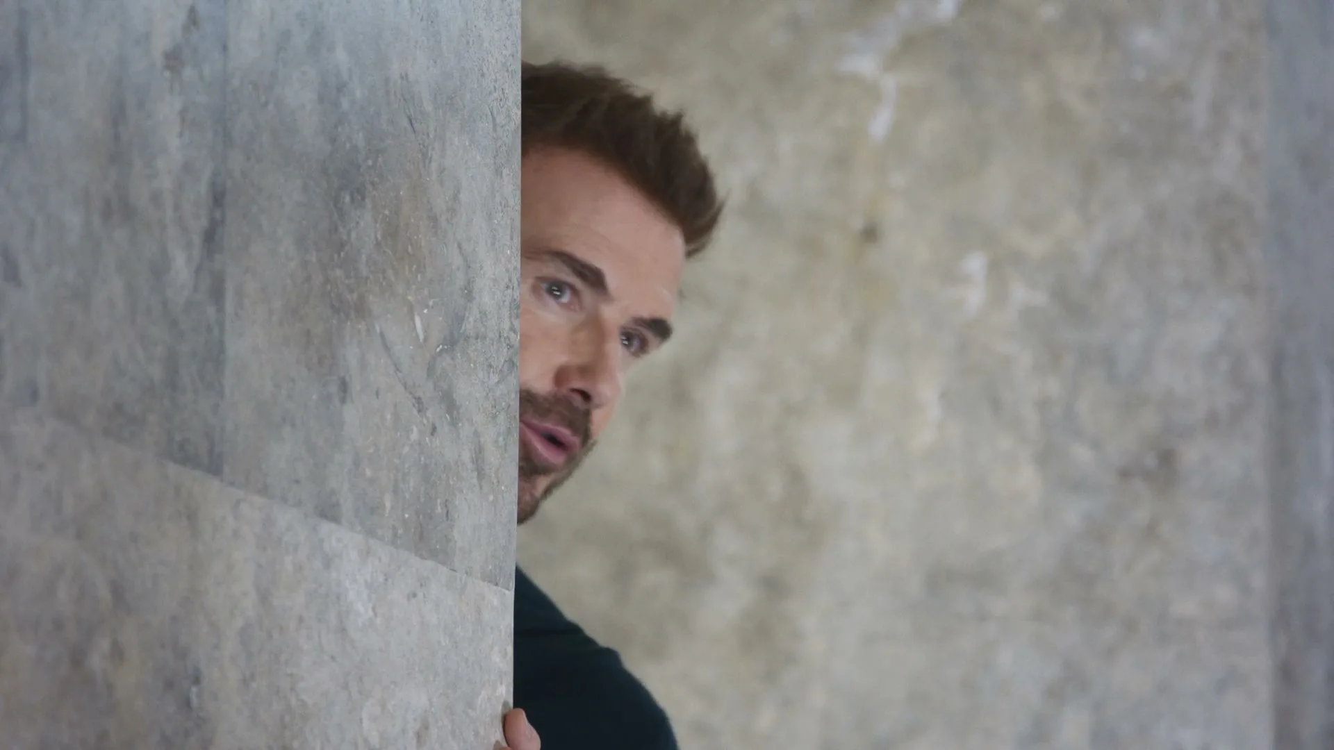 A man with a beard and dark shirt peeks from behind a stone wall, looking to the right with a curious expression.