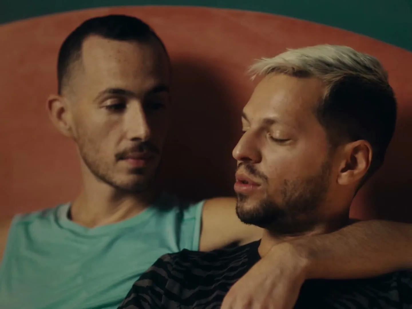 Two men, one with bleached hair and a black shirt, the other with a teal shirt, embrace and talk intimately on a couch in a dimly lit room.