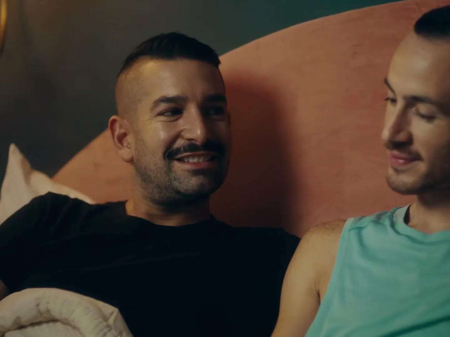 Two men, one with a mustache and black t-shirt, the other in a teal tank top, lie in bed, smiling and looking at each other.