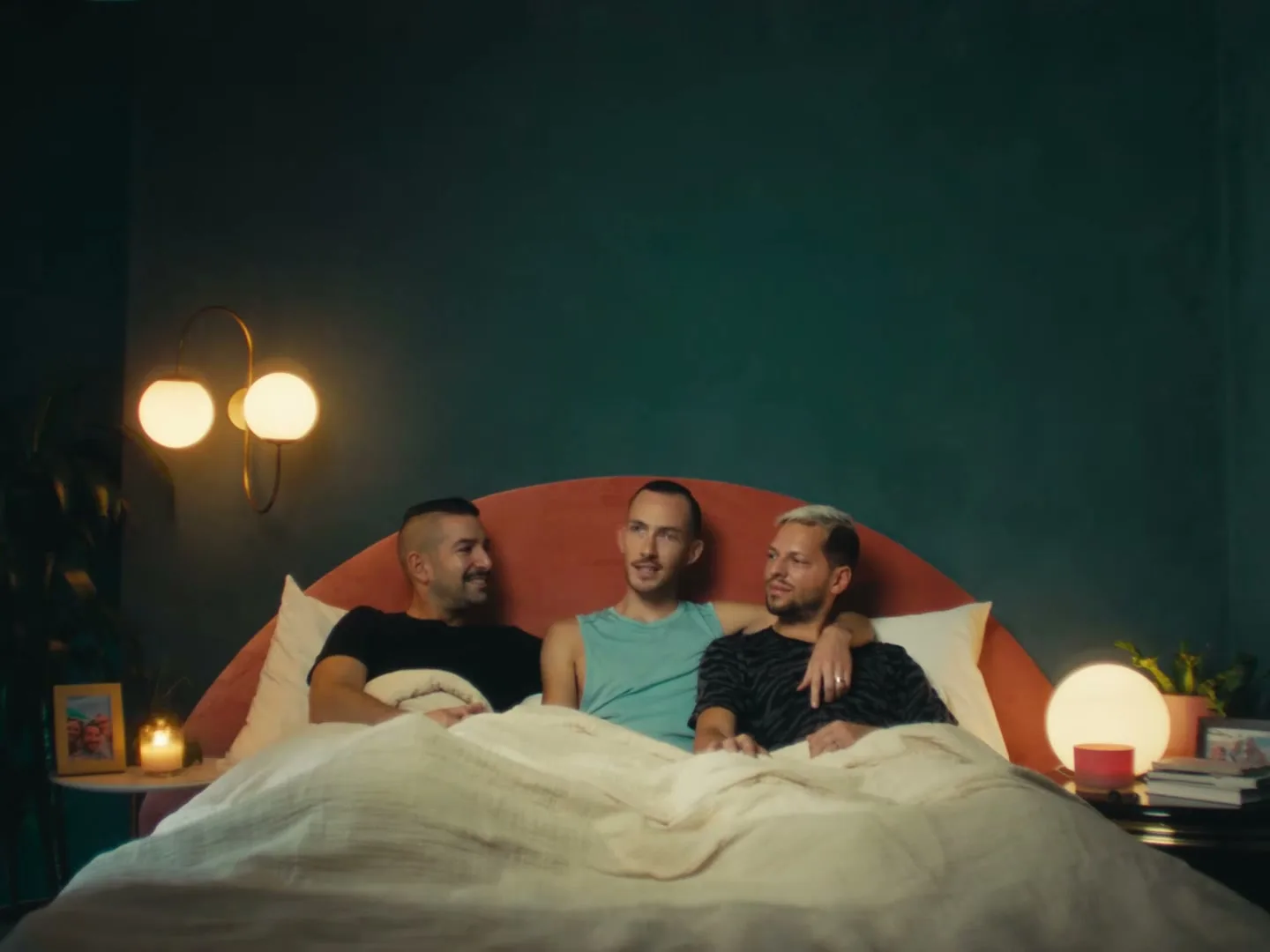 Three men, two with dark hair and one with blonde, lie in bed together, talking and smiling in a cozy bedroom.