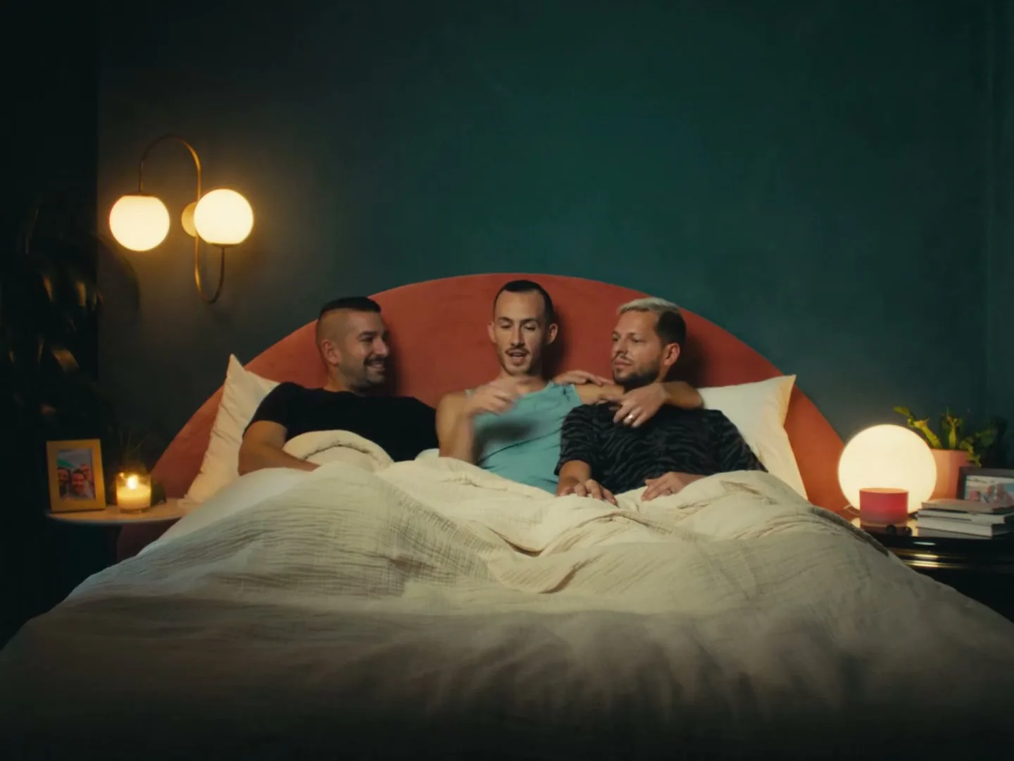 Three men lie in bed, covered by a duvet, talking and smiling in a cozy bedroom at night.