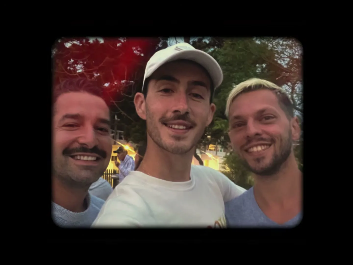 Three men, two with mustaches and one with a cap, smile at the camera in a selfie-style shot, with trees and people in the background.