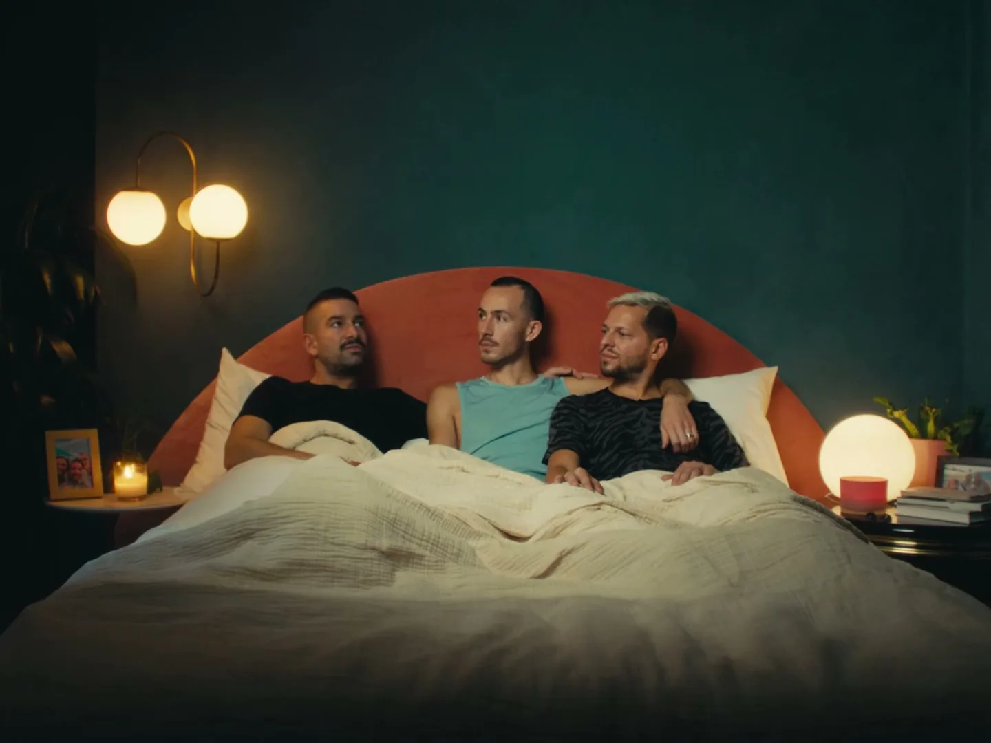 Three men lie in bed, two of them looking at the third, who is speaking. They are in a cozy bedroom with warm lighting.
