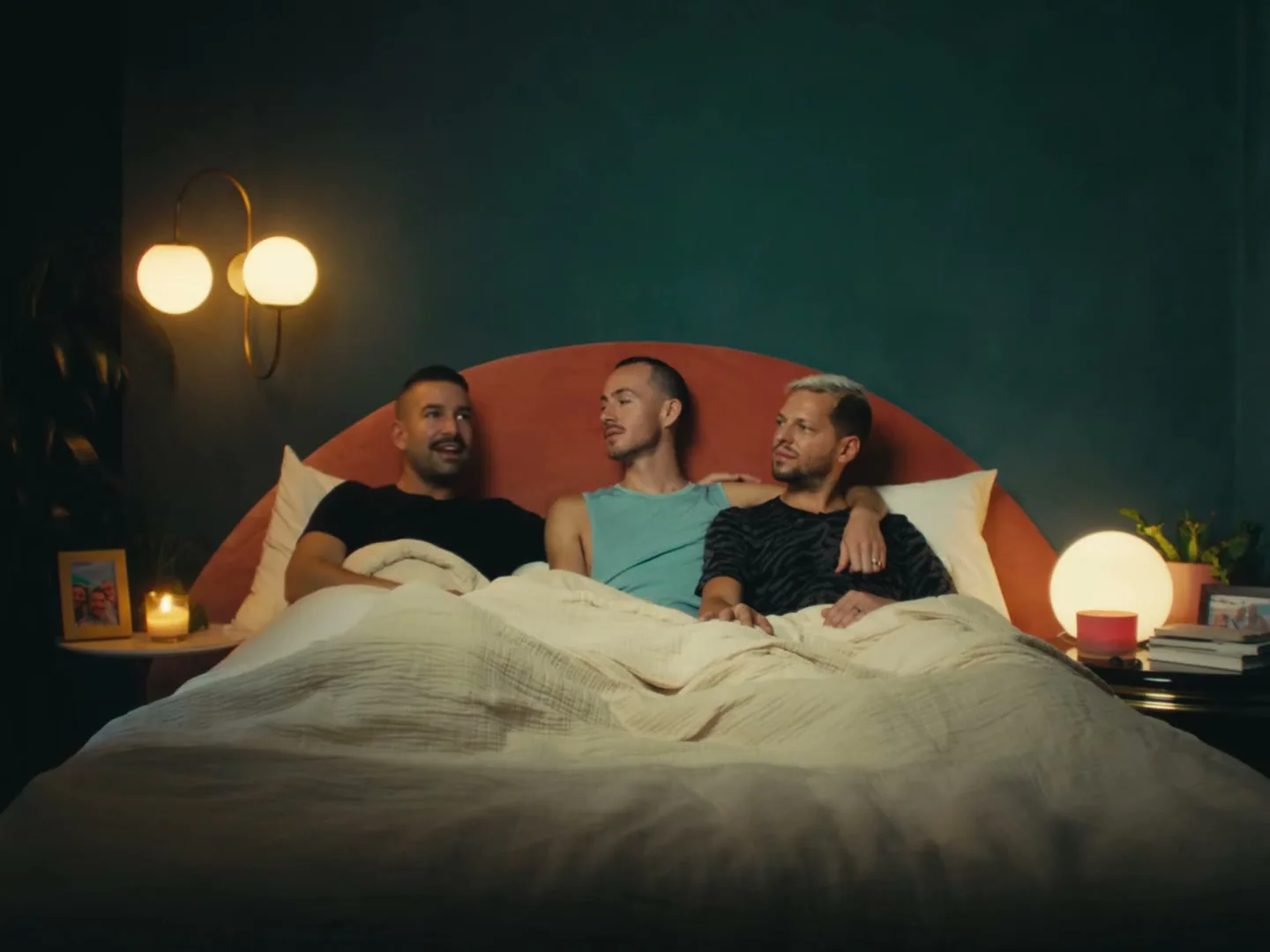 Three men lie in bed, two looking at the third, who smiles, in a cozy bedroom at night.