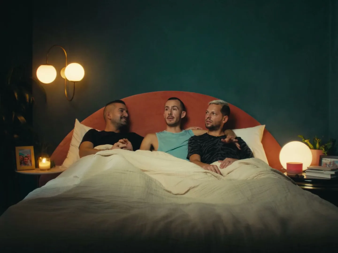 Three men lie in bed, two of them with arms around each other, talking in a cozy bedroom at night.
