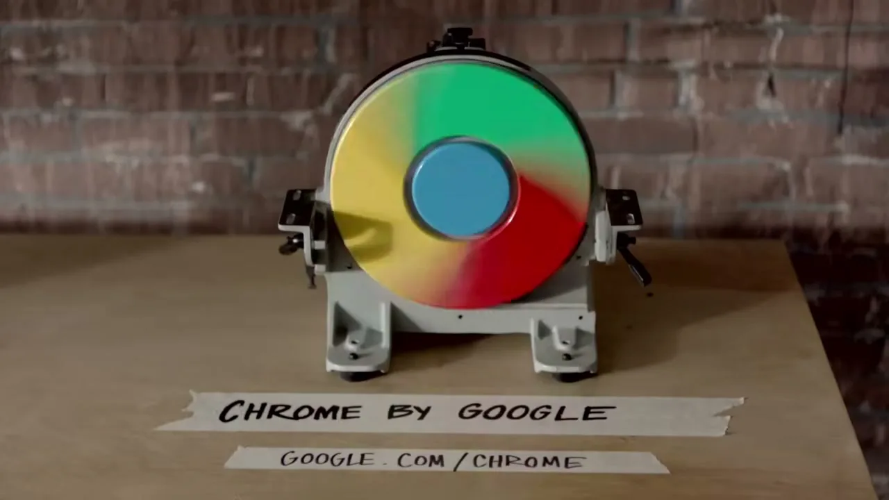 A Google Chrome logo, made of a spinning, colorful disc, is mounted on a small, grey machine on a wooden surface, with a brick wall in the background.