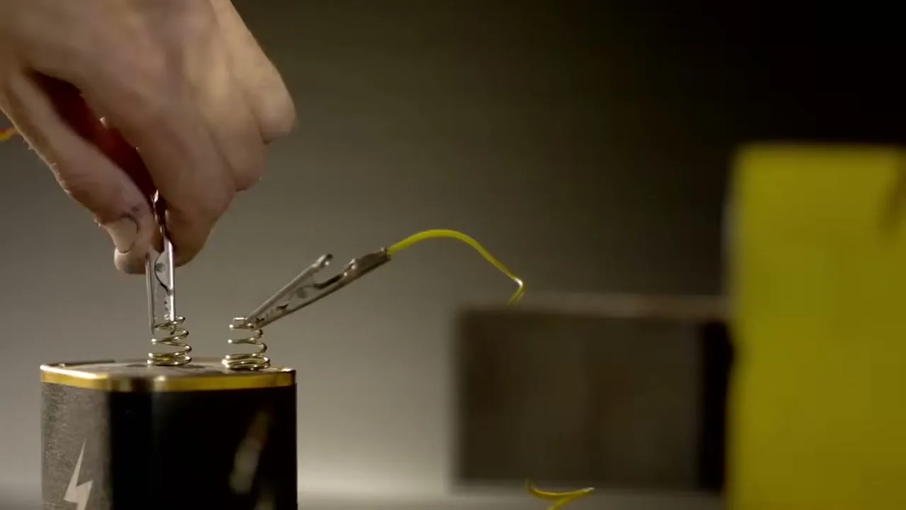 A person's hand connects a yellow wire to a battery with alligator clips, demonstrating an electrical circuit in a dimly lit setting.