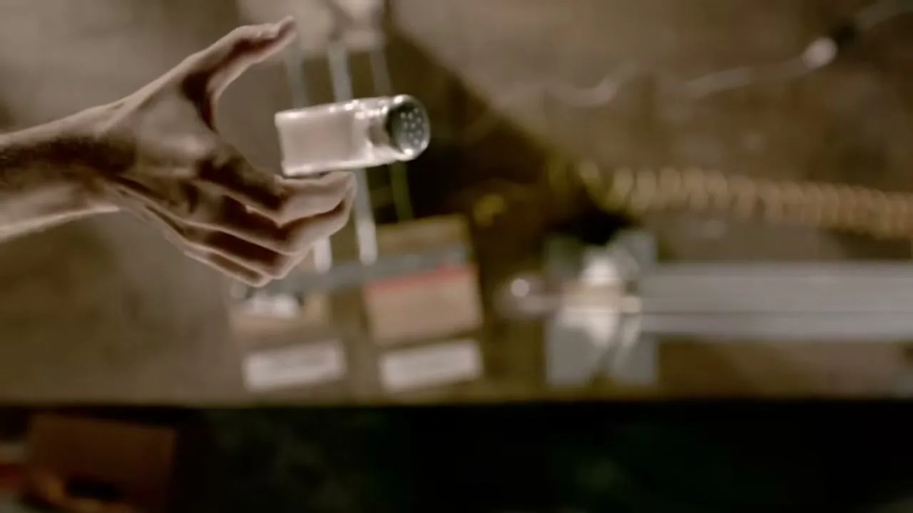 A male hand holds a salt shaker over a wooden table with various objects, including a lightbulb and wires, in a dimly lit room.