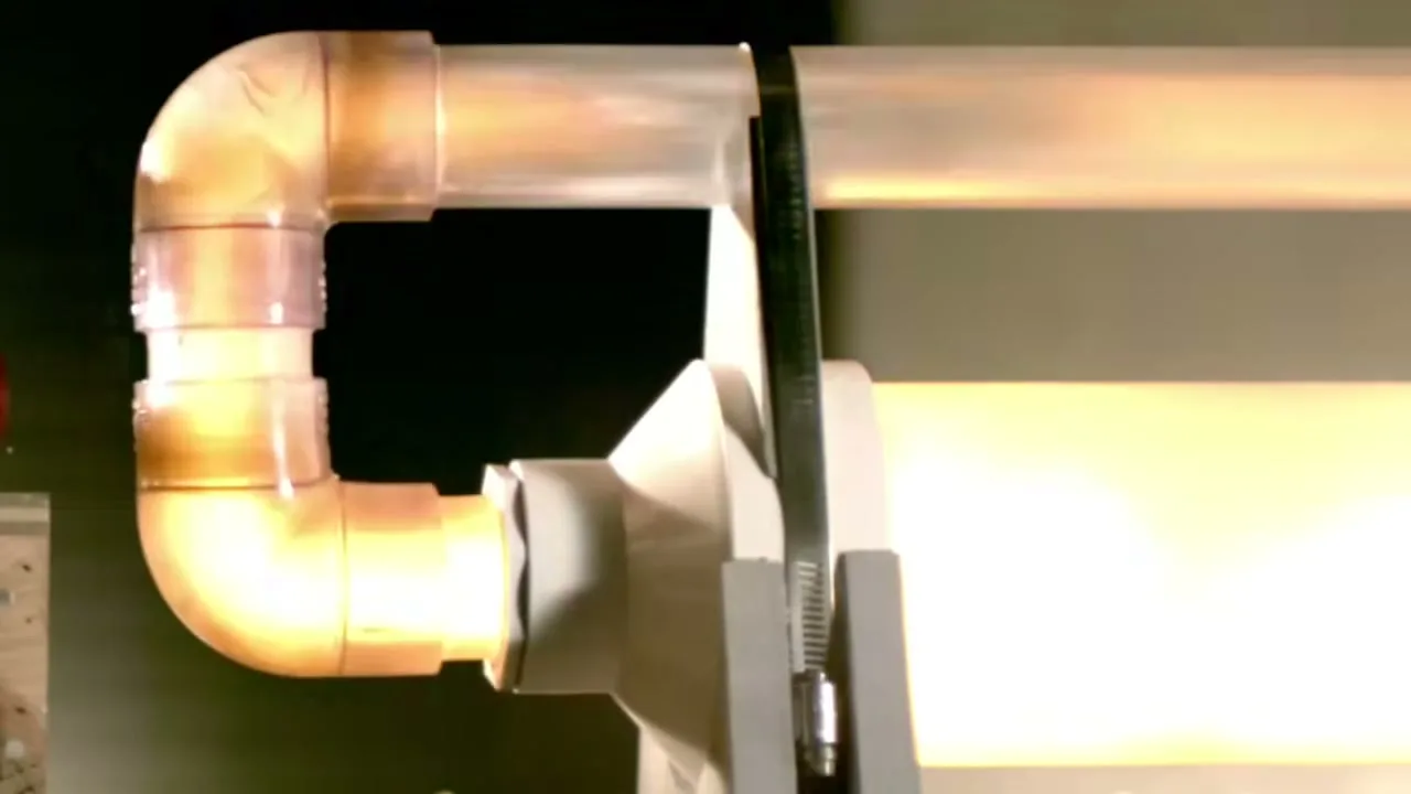 A close-up shot shows a clear pipe with glowing orange liquid flowing through it, connected to a white mechanism, in a dark setting.
