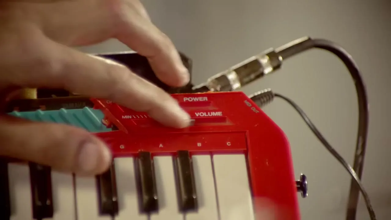 A person's hand adjusts the volume slider on a small red and white electronic keyboard, with cables connected.