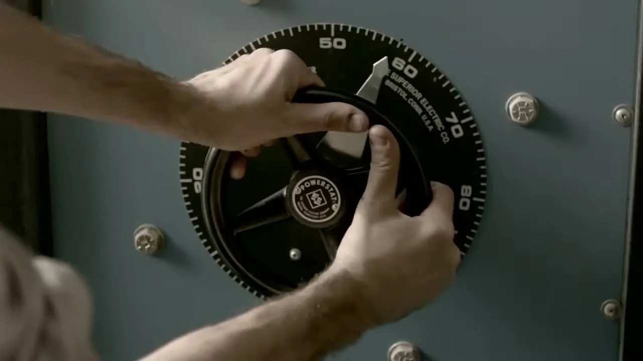 A man's hands adjust a large, black dial on a blue-grey industrial machine, turning it clockwise in a close-up shot.