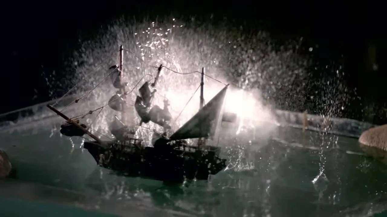 A miniature pirate ship with black sails and a skull and crossbones flag is hit by a sparkler and water splashes around it in a dark setting.
