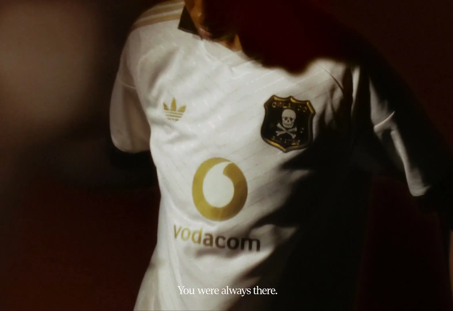A person wearing a white soccer jersey with a skull and crossbones emblem and "Vodacom" logo is shown in a dimly lit setting.