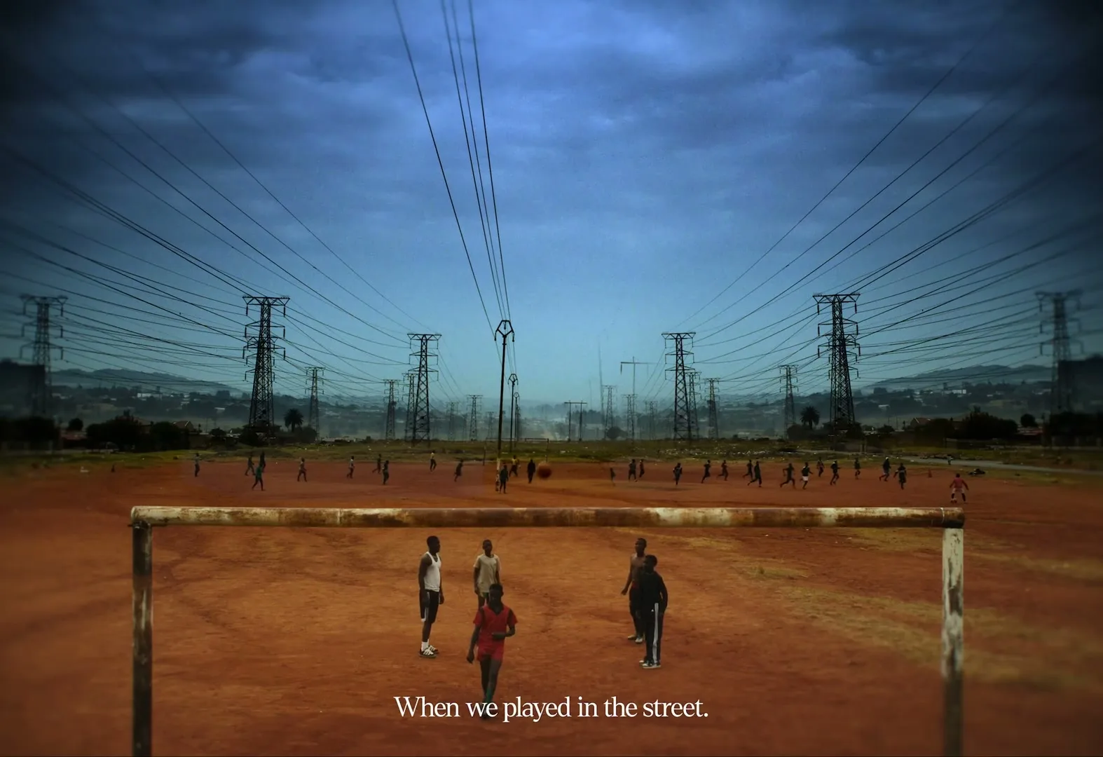 A wide shot shows a group of children playing soccer on a dusty field under a dramatic sky, with power lines and towers in the background.