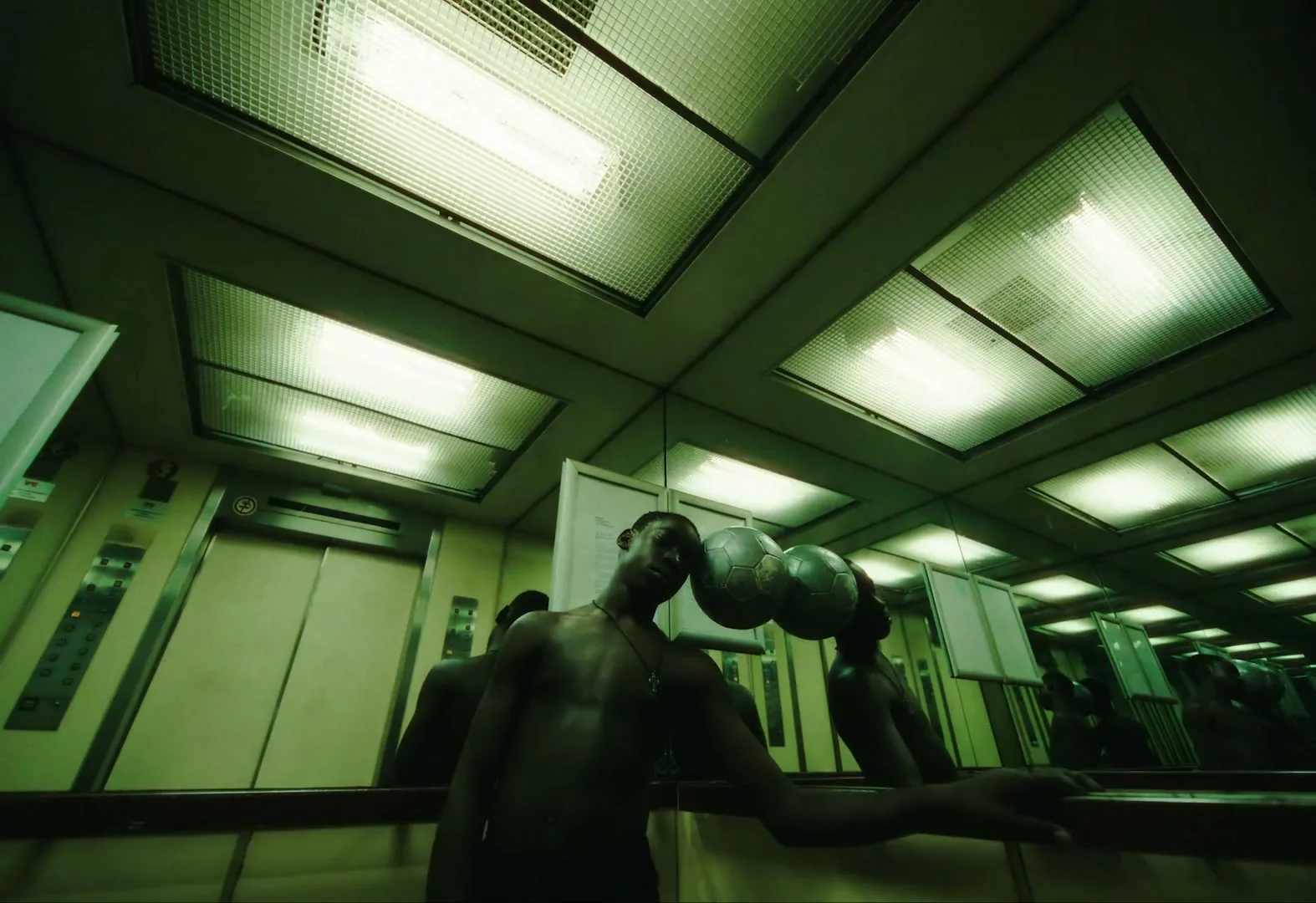 A shirtless young man balances a silver soccer ball on his head in a mirrored elevator, looking directly at the camera.