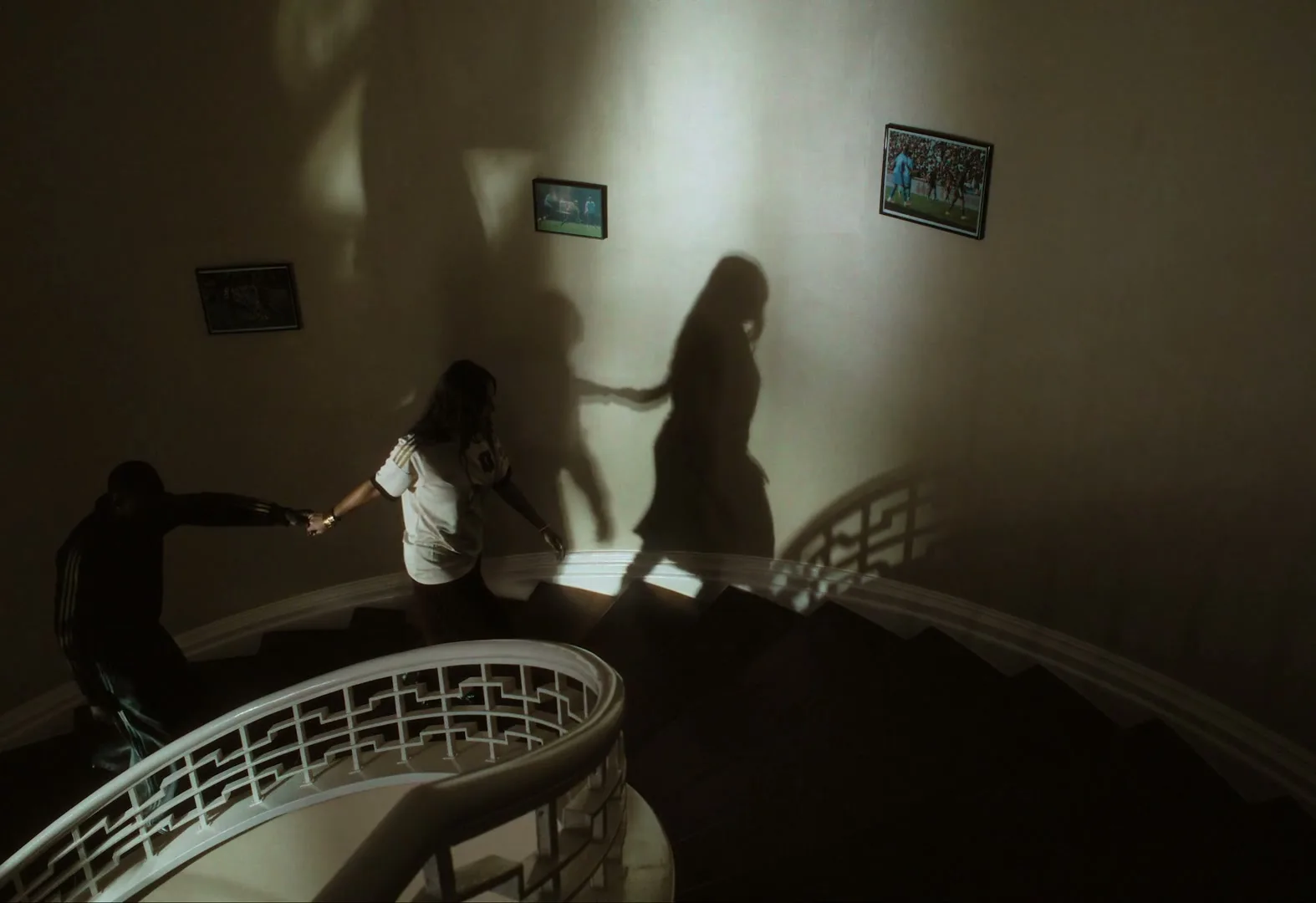 Two people, a man and a woman, walk up a dark, winding staircase, holding hands, with their shadows cast large on the wall.