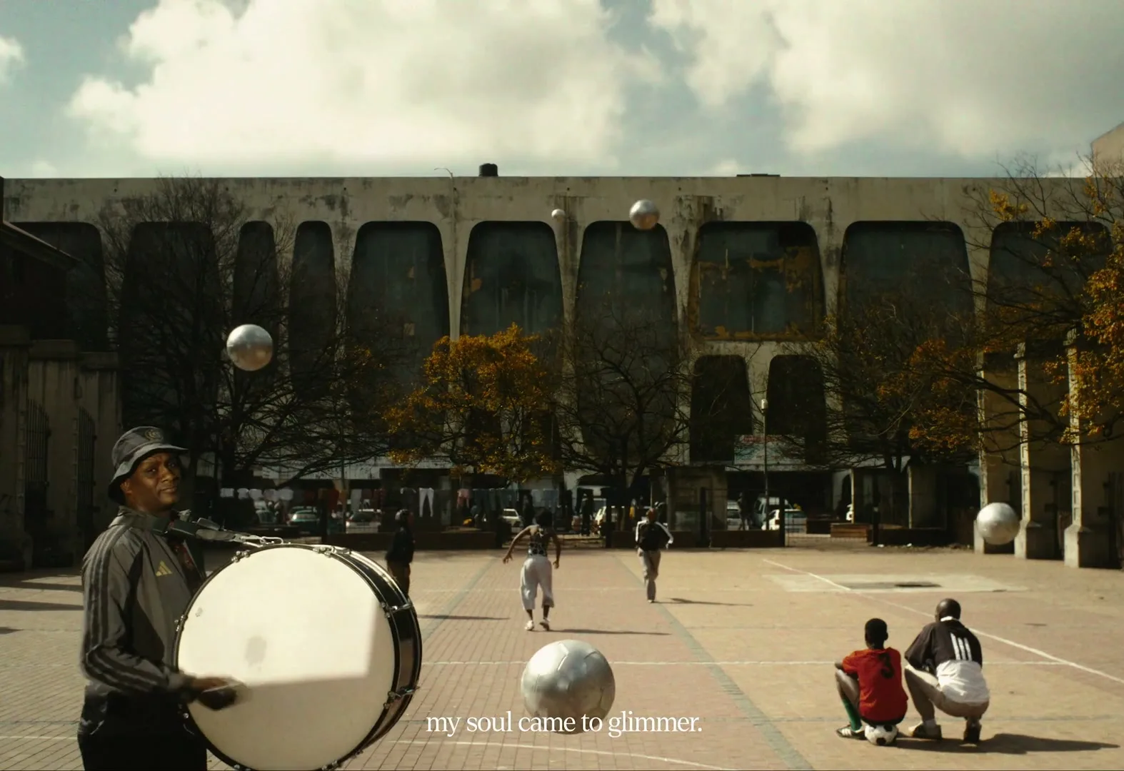 A man in a bucket hat plays a bass drum while children play soccer in a paved courtyard with a large concrete building in the background.