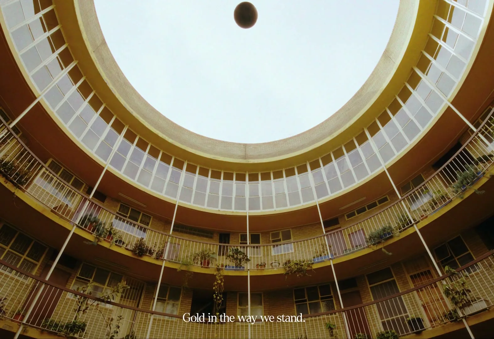A wide shot of a circular apartment building courtyard with balconies and windows, looking up at a bright sky, with a dark sphere floating in the center.