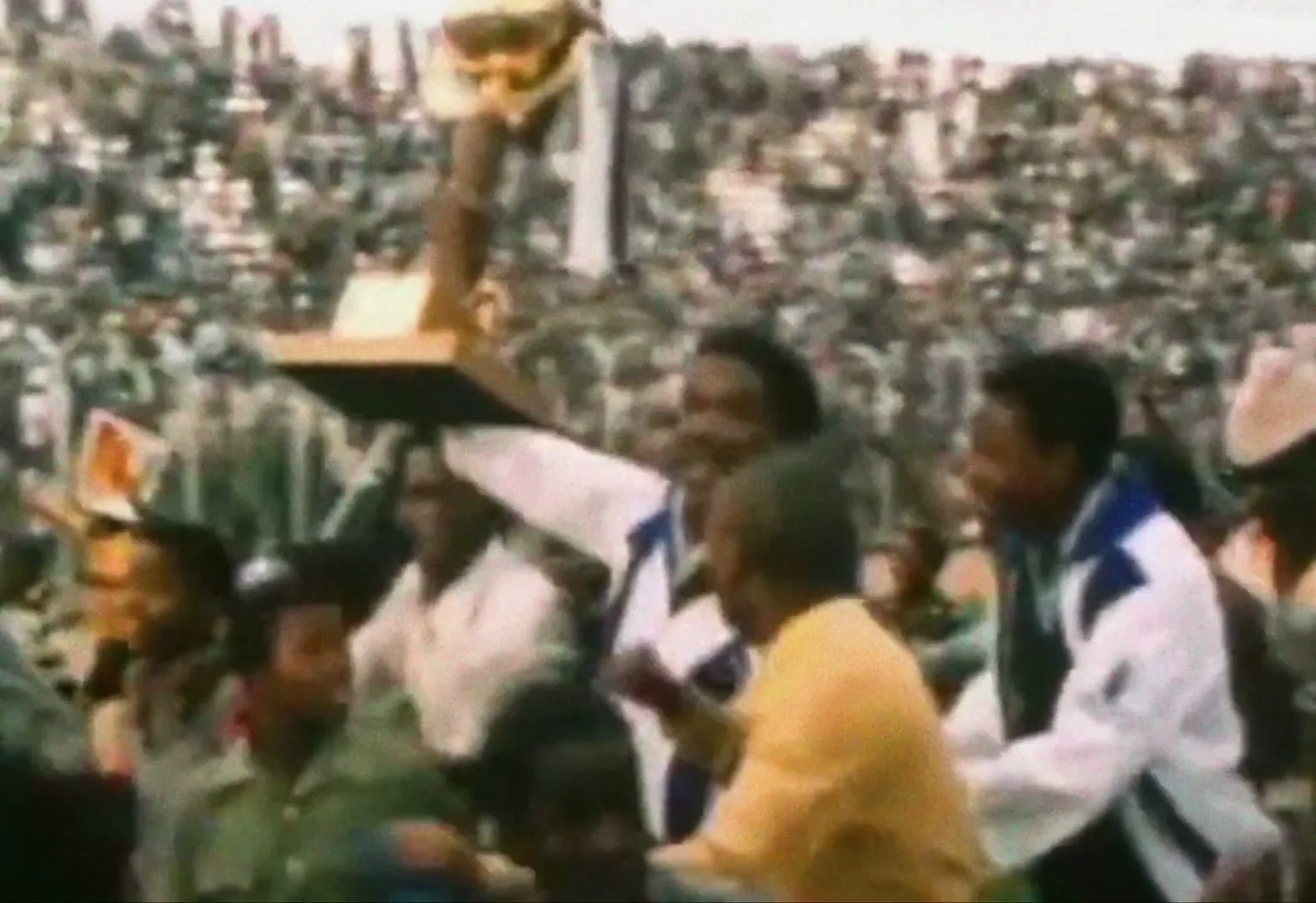 A crowd of people celebrate as a man in a white and blue jacket holds a trophy aloft in a stadium.