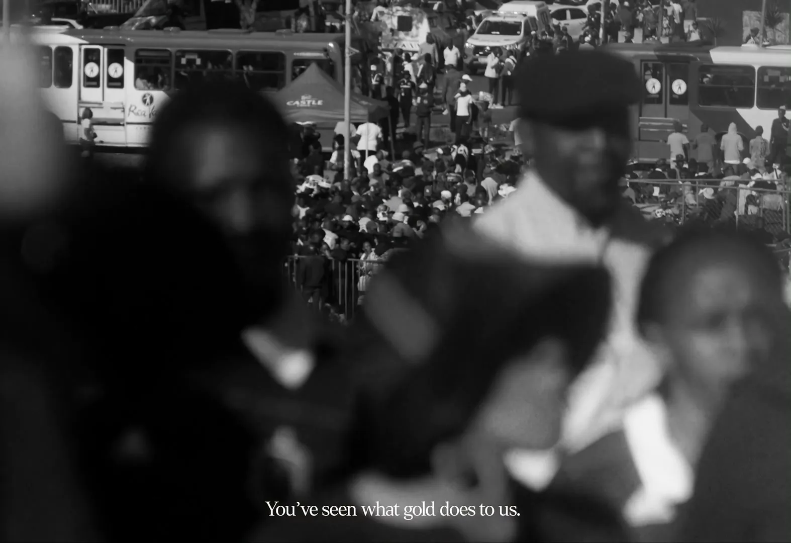 A black and white, blurry shot of a crowd of people, with three men in the foreground out of focus, and a bus and cars in the background.