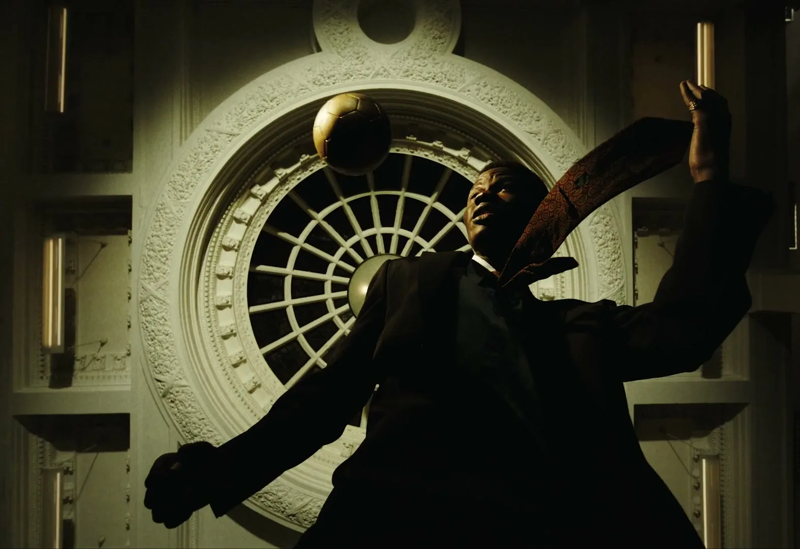 A man in a suit juggles a golden soccer ball in front of a large, ornate circular window in a grand building.