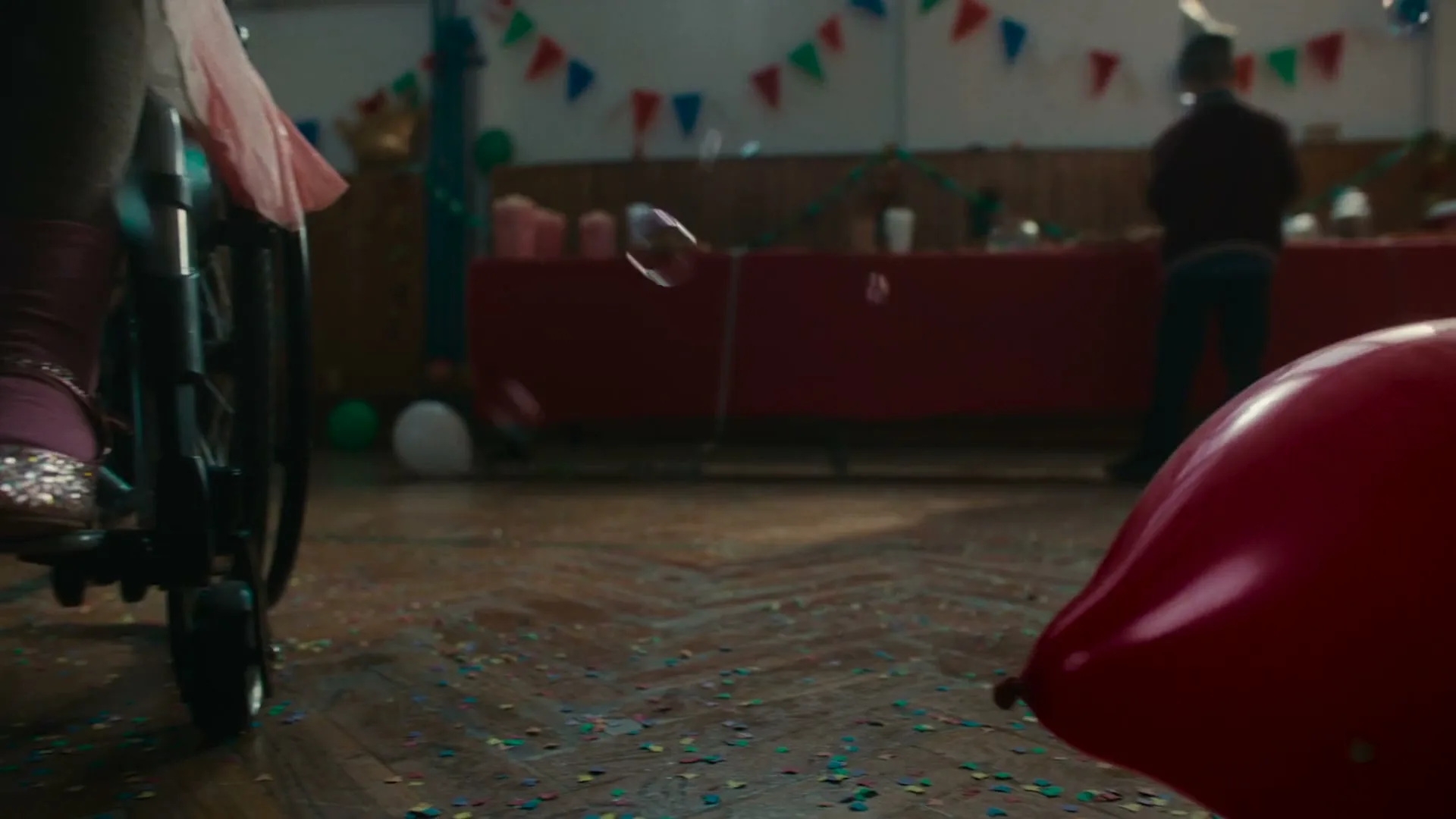 A low-angle shot captures the feet of children dancing and running around a girl in a wheelchair at a lively birthday party in a school gym.