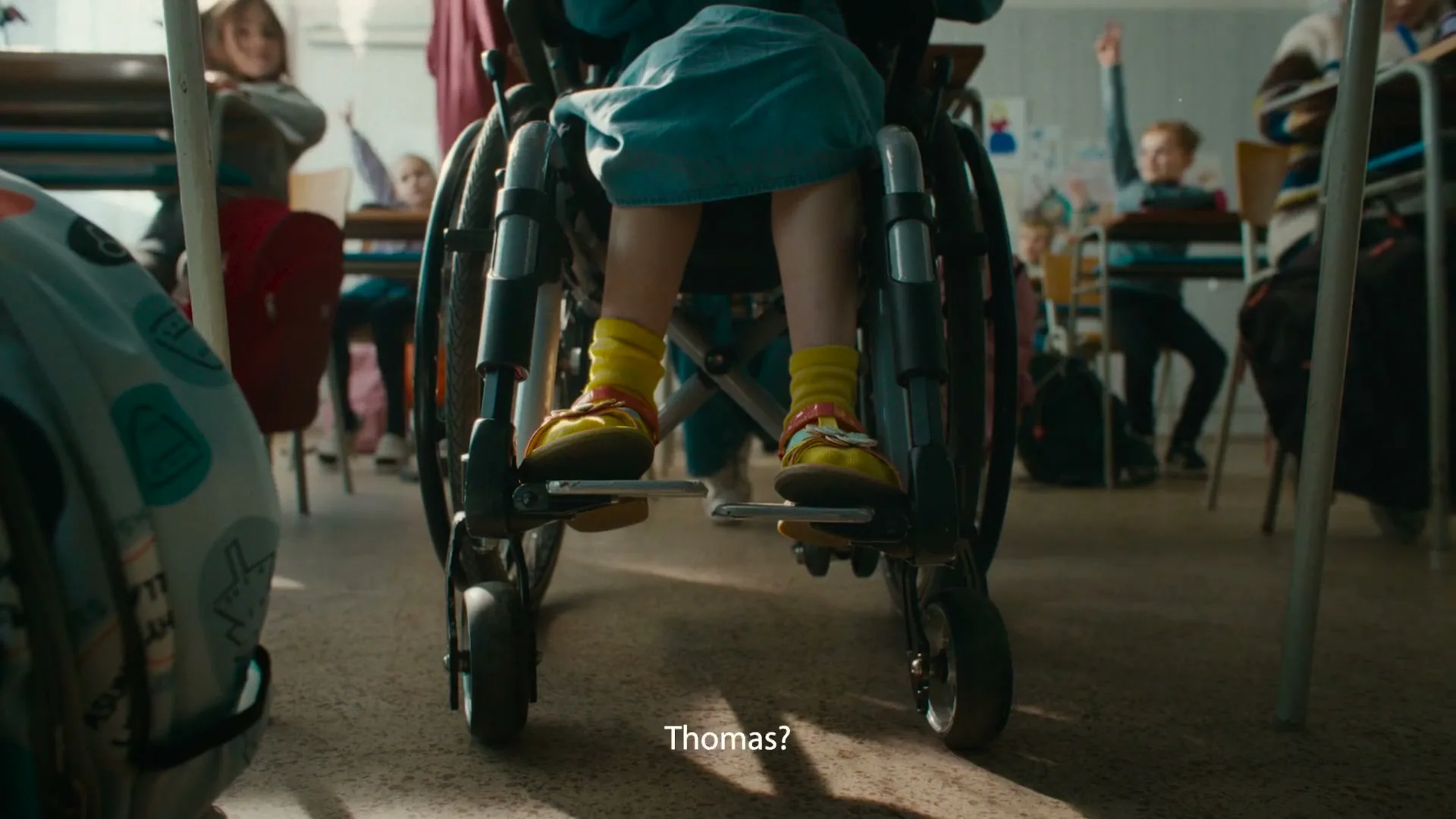 A low-angle shot shows a child in a wheelchair from behind, wearing a blue dress, yellow socks, and sandals, in a classroom with other children.