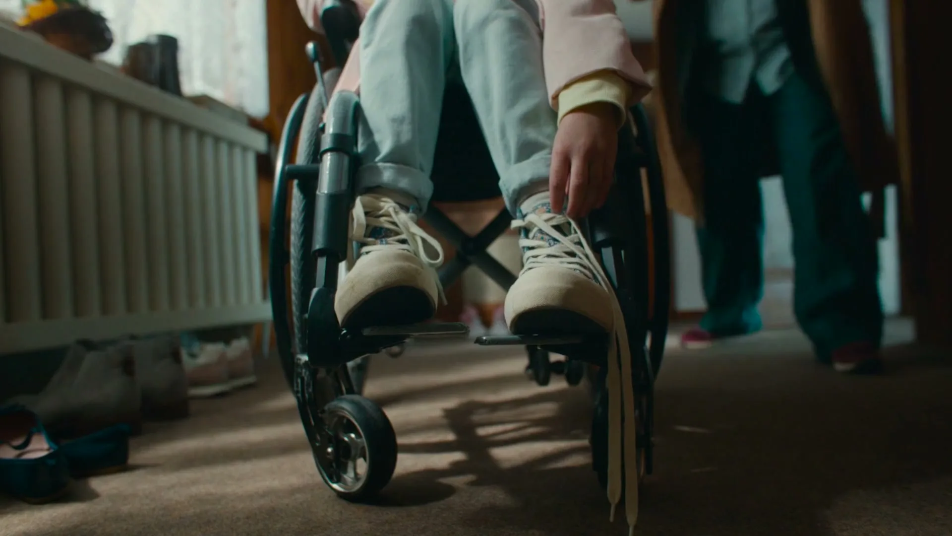 A person in a wheelchair has their shoelaces tied by another person, in a warm, inviting interior setting.