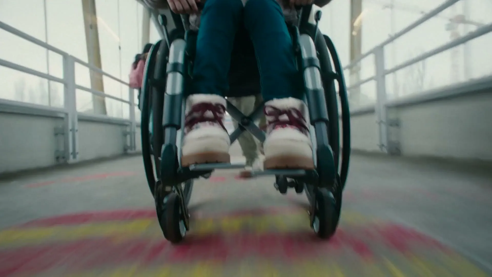 A person in a wheelchair, wearing blue jeans and white furry boots with red laces, moves forward on a bridge with colorful chalk drawings on the ground.