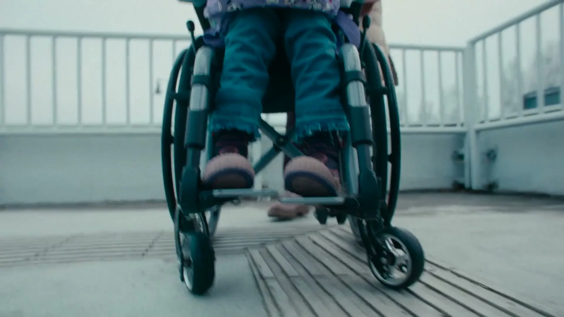 A child in a wheelchair is pushed forward by an adult, showing only their legs and the wheelchair on a concrete path with a railing.