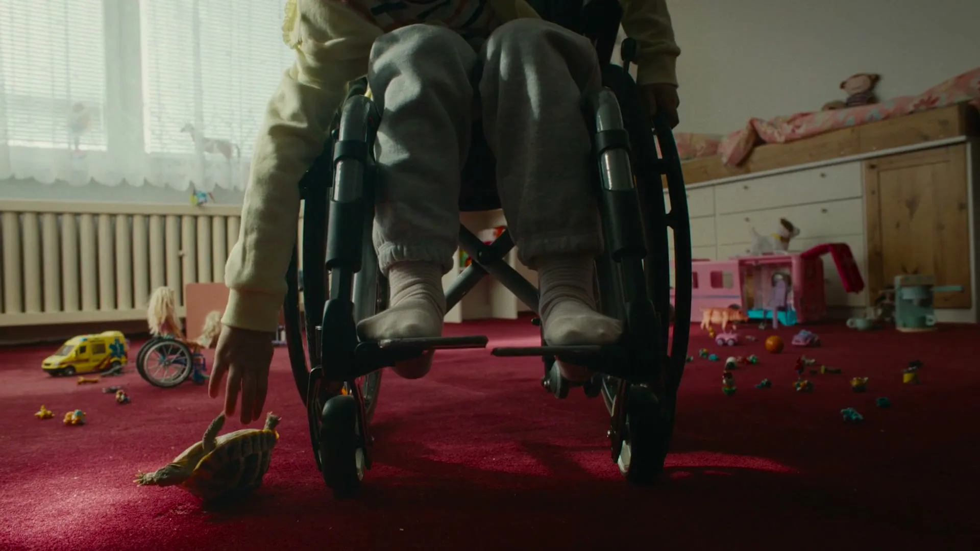 A child in a wheelchair reaches down to flip over a toy turtle on a red carpet in a messy bedroom.
