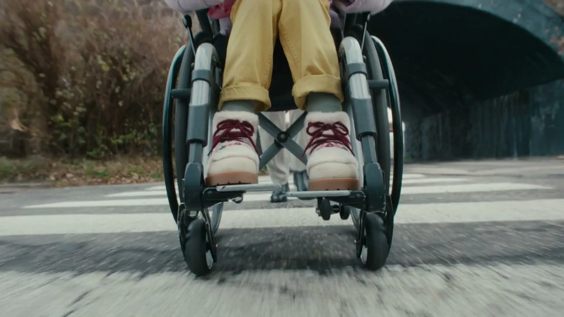 A low-angle shot shows a person's feet in a wheelchair crossing a street, with a car passing by in the background.
